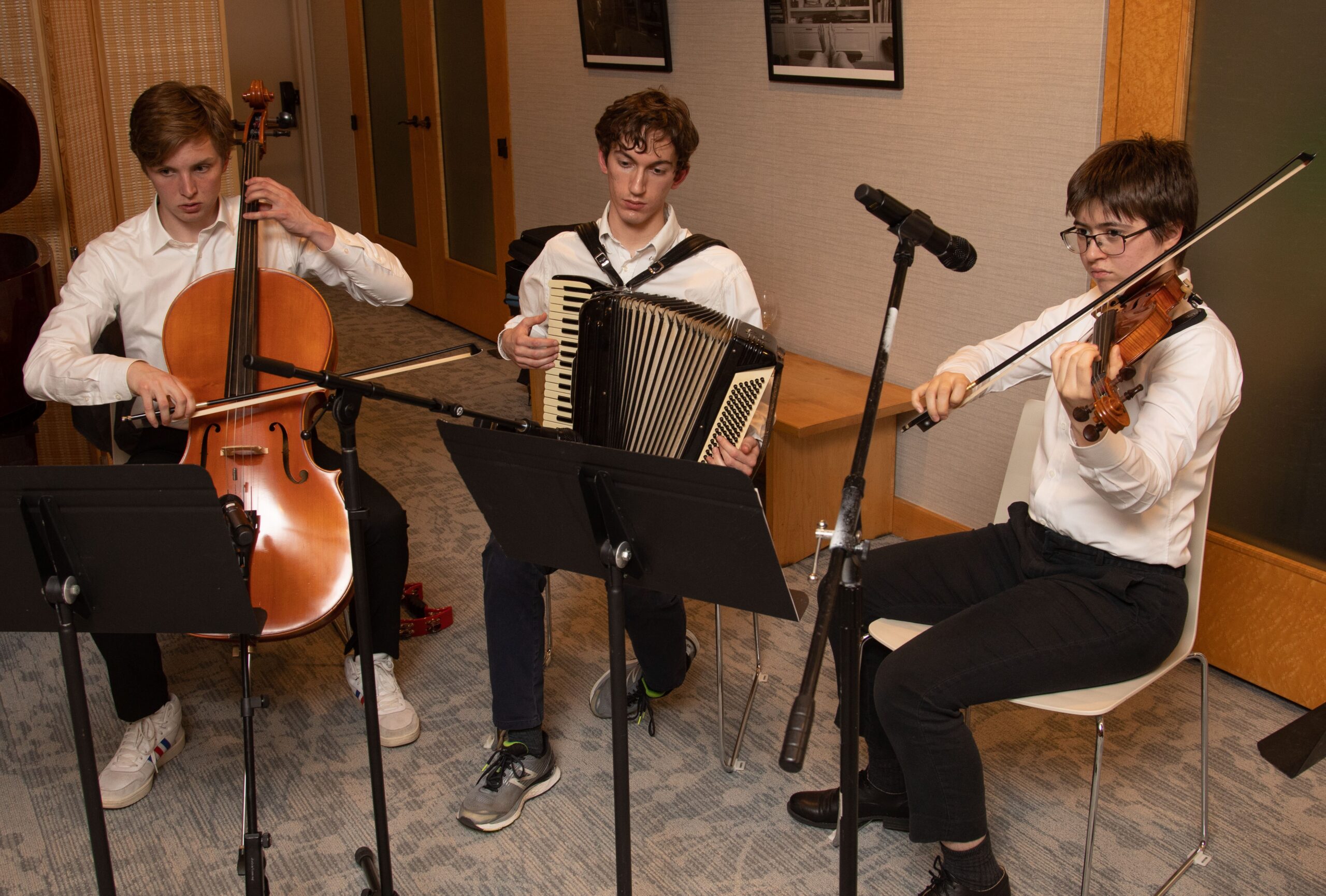 Three people sitting together playing klezmer music, one with a cello, one with an accordion and one with a violin. All are dressed formally in white shirts and black pants.