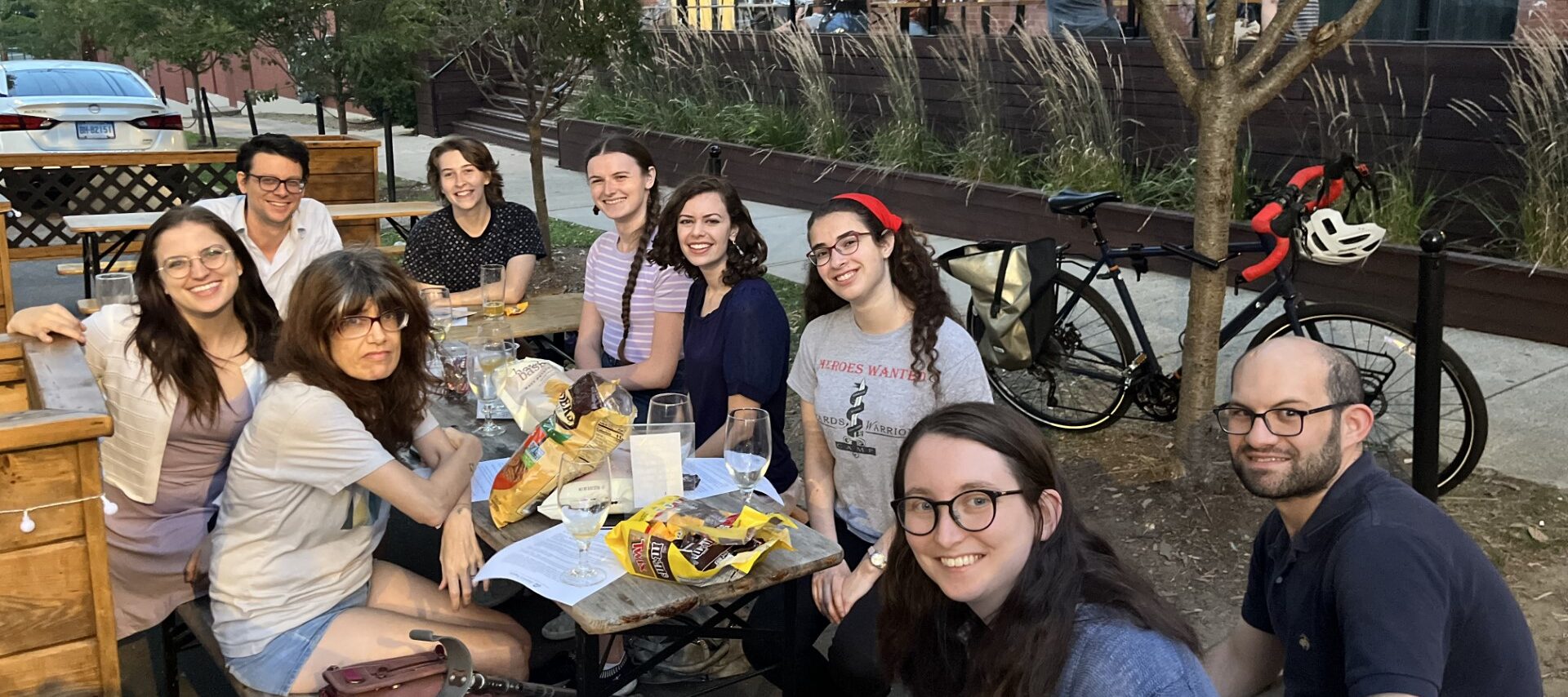 People sit around a table outside, smiling at the camera. On the table are snacks and beers.